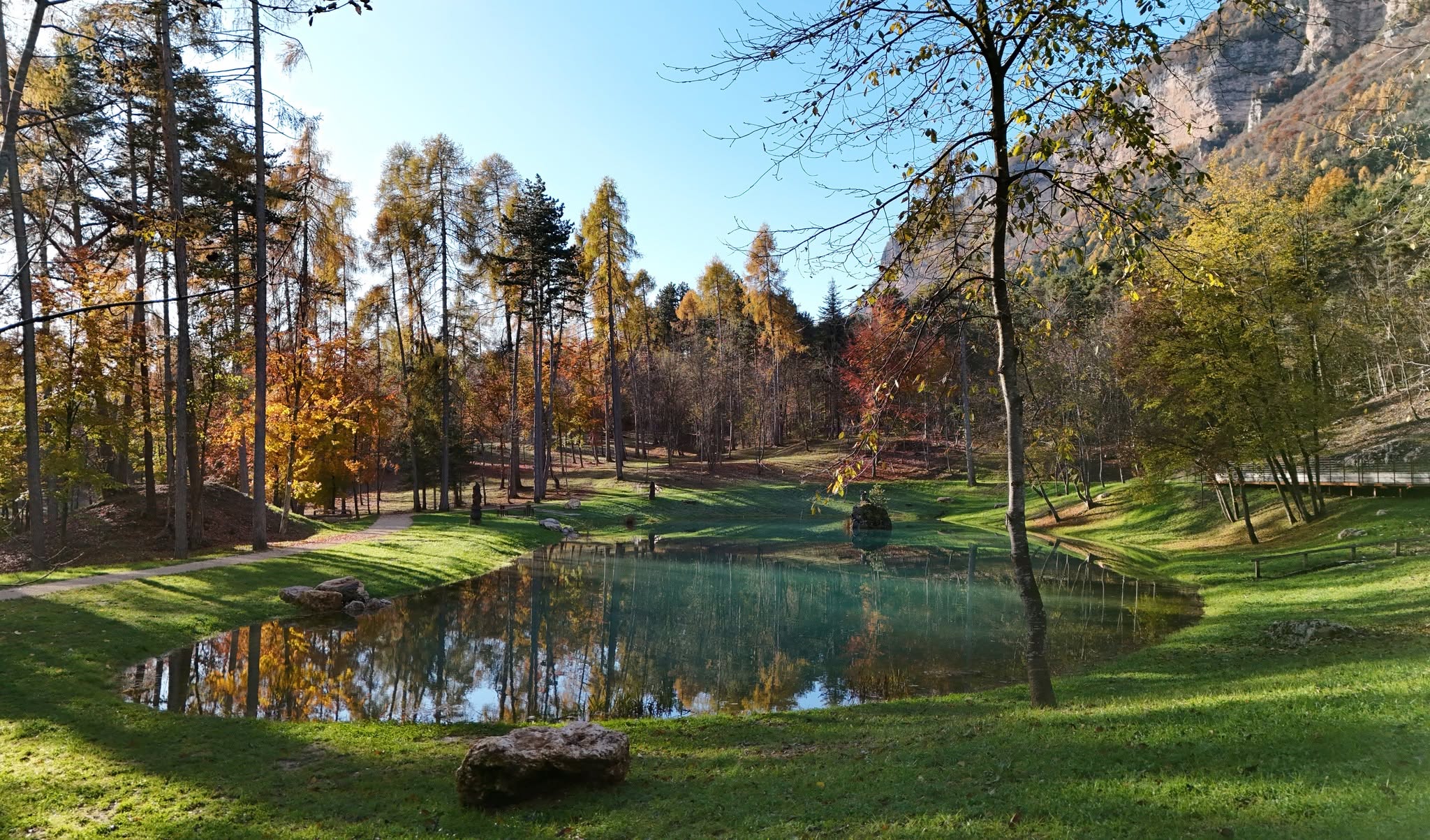 Specchio d'acqua e prato alberato in autunno, con la parete rocciosa sullo sfondo: un intervento di paesaggio dello studio AMP